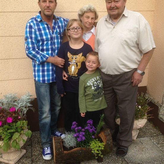 Eine vierköpfige Familie steht gemeinsam vor einer Wand mit Blumen. Der Mann links trägt ein blau kariertes Hemd, während die Frau in der Mitte ein orangefarbenes Oberteil trägt. Der Junge vorne trägt ein grünes Shirt mit Katzendesign.
