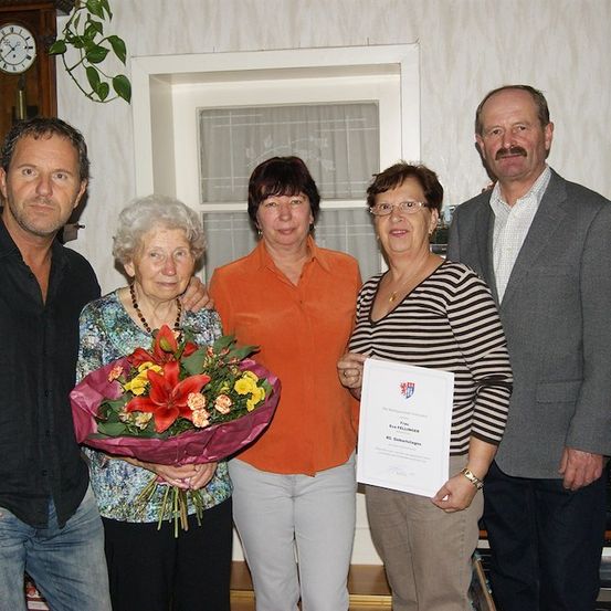 Eine Gruppe von fünf Personen steht lächelnd für ein Foto in einem Raum. Eine Frau in der Mitte hält einen Blumenstrauß und ein Zertifikat. Die Frau links trägt eine Halskette. Hinter ihnen befindet sich ein Fenster mit Vorhängen, einer Uhr und einer Pflanze.