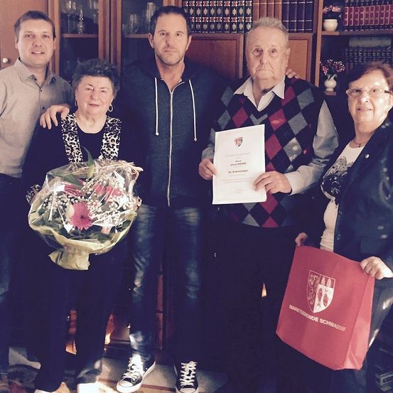 Ein Familienfoto mit einem Mann, der ein Zertifikat und einen Blumenstrauß hält, steht mit drei anderen Personen in einem Raum mit Bücherregalen.