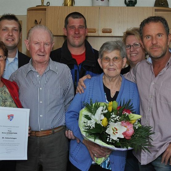 Eine Gruppe von Menschen versammelt sich zu einer Feier. Eine ältere Frau hält einen Blumenstrauß und ein Zertifikat. Der Mann neben ihr hält ein Zertifikat. Hinter ihnen lächeln eine Frau mit Brille und ein Mann mit Bart. Im Hintergrund befinden sich Holzschränke mit Gegenständen obenauf.