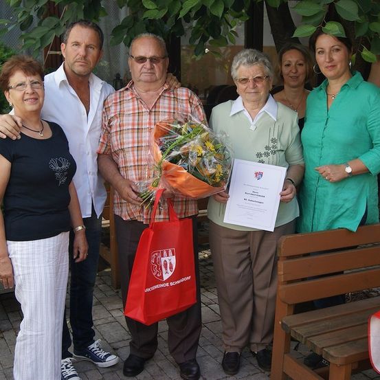 Eine Gruppe von Menschen steht draußen, einer hält einen Blumenstrauß und ein Zertifikat, mit einer roten Tasche und einer Holzbank im Hintergrund.