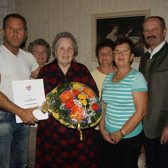 Eine Gruppe von Menschen versammelt sich zu einer Feier, wobei ein Mann ein Zertifikat und einen Blumenstrauß hält. Eine ältere Frau steht in der Mitte, lächelt und ist von anderen Frauen umgeben. Ein gerahmtes Bild hängt an der Wand hinter ihnen.