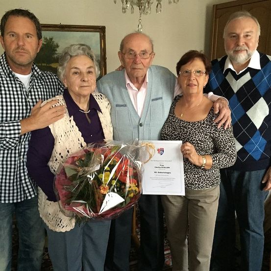 Eine Gruppe von fünf Personen, darunter eine ältere Frau, die einen Blumenstrauß und ein Zertifikat in der Hand hält, posiert für ein Foto in einem Raum.