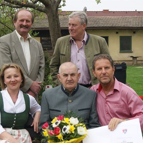 Fünf Personen posieren für ein Foto vor einem Haus. Der Mann in der Mitte hält ein Zertifikat und einen Blumenstrauß. Die Frau auf der linken Seite trägt ein traditionelles Kleid. Der Mann auf der rechten Seite trägt ein rosa Hemd. Hinter ihnen steht ein Haus mit einem roten Dach und einem Baum.