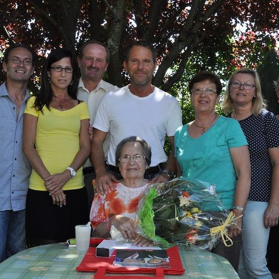 Eine Gruppe von Menschen, einschließlich einer älteren Frau, versammelt sich um einen Tisch mit einem Blumenstrauß und einem Geschenk. Sie stehen unter einem Baum mit roten Blättern.