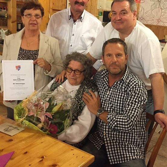 Eine Gruppe von Menschen versammelt sich um eine Frau, die ein Zertifikat und Blumen hält. Die Frau sitzt und wird von einem Mann in einem karierten Hemd umarmt.