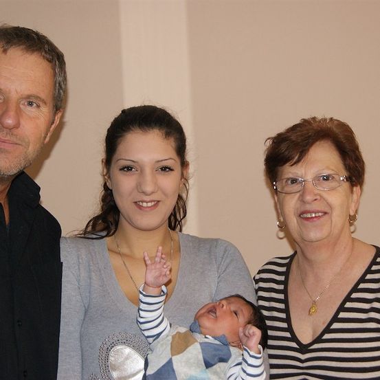 Ein Familienfoto mit einem lächelnden Mann, einer Frau mit einem Baby und einer älteren Frau mit Brille. Das Baby macht ein Friedenszeichen.