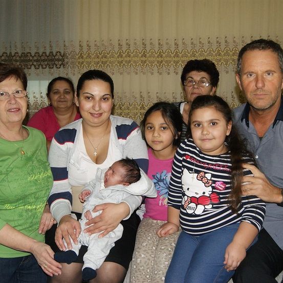 Ein Familienfoto mit einer Frau, die ein Neugeborenes hält, umgeben von anderen Familienmitgliedern in einem Raum mit einem Vorhang im Hintergrund.