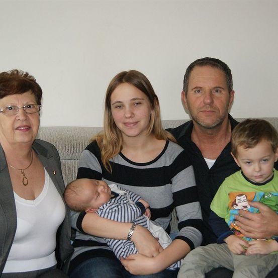 Ein Familienfoto mit einer lächelnden Frau, die ein Baby hält, einem Mädchen mit einem schlafenden Baby, einem Mann und einem Jungen, der in die Kamera schaut.