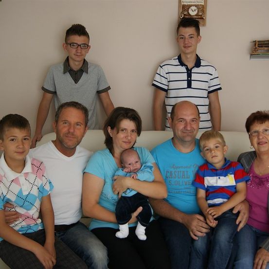 Ein Familienfoto mit einem Neugeborenen. Die Familie sitzt auf einer Couch, zwei Erwachsene stehen hinter ihnen. Das Baby wird von einer Frau in einem blauen Shirt gehalten.