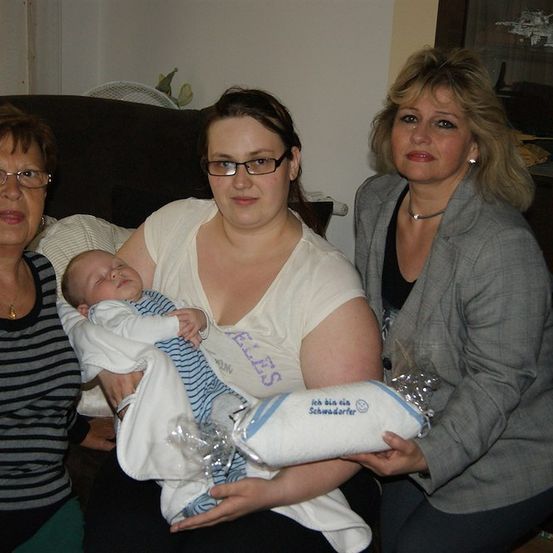 Drei Frauen posieren für ein Foto mit einem Baby. Das Baby schläft in den Armen der Frau in der Mitte. Die Frau auf der linken Seite trägt eine Brille und eine Halskette. Die Frau auf der rechten Seite trägt einen Blazer und hält ein eingewickeltes Geschenk.