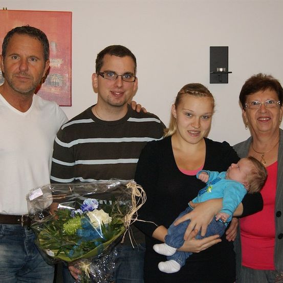 Eine vierköpfige Familie steht eng beieinander und lächelt. Ein Mann hält einen Blumenstrauß, eine Frau ein Baby, und zwei ältere Erwachsene stehen neben ihnen. Sie befinden sich in einem Raum mit einer weißen Wand und einem gerahmten Bild.