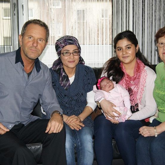 Eine Familie aus vier Personen, bestehend aus einem Mann, zwei Frauen und einem Baby, sitzt auf einem Sofa. Das Baby wird von einer Frau gehalten, die einen rosa Schal trägt. Der Mann trägt ein graues Hemd und die andere Frau ein grünes Hemd. Sie alle lächeln und scheinen für ein Foto zu posieren.