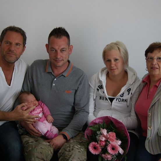 Eine Familie von vier Personen sitzt zusammen, ein Mann hält ein Baby, eine Frau hält einen Blumenstrauß und zwei Frauen lächeln in die Kamera.