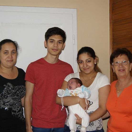 Eine vierköpfige Familie posiert für ein Foto. Die Frau links trägt ein schwarzes Shirt mit Katzendesign. Der Mann in der Mitte trägt ein rotes Shirt. Die Frau rechts trägt ein orangefarbenes Shirt und hält ein Baby. Das Baby trägt ein weißes Outfit.