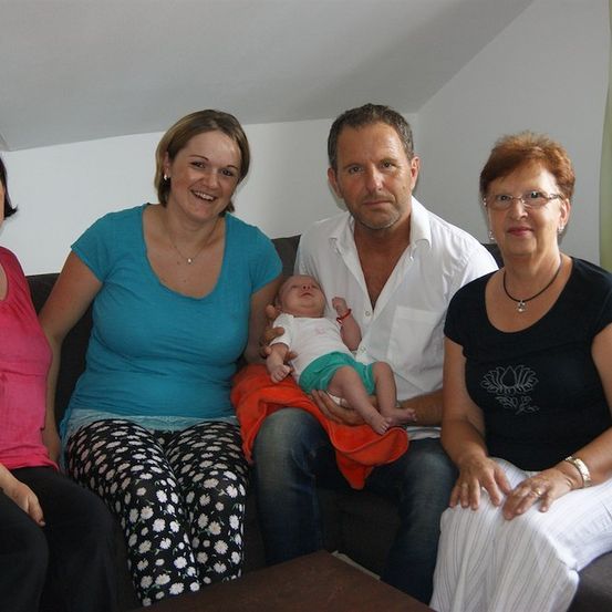 Eine Familie mit vier Personen, bestehend aus einem Mann, einer Frau und einem Baby, sitzt auf einem Sofa und lächelt für ein Foto.