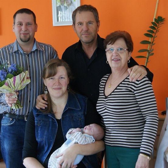 Eine vierköpfige Familie, einschließlich eines Neugeborenen, steht in einem Raum mit orangefarbener Wand zusammen. Der Mann auf der linken Seite hält einen Blumenstrauß, und die Frau auf der rechten Seite hält das Baby. Der Mann in der Mitte lächelt und hat seinen Arm um die Frau auf der rechten Seite gelegt.