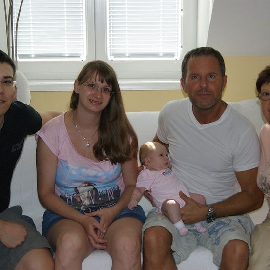 Eine vierköpfige Familie sitzt auf einer Couch, wobei ein Mann ein Baby hält, eine Frau in der Mitte eine Brille trägt und zwei weitere auf beiden Seiten sitzen.