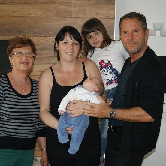 Eine vierköpfige Familie, einschließlich eines Babys, steht in einer Küche zusammen. Die Frau hält das Baby, während ein Mann und ein Mädchen auf beiden Seiten stehen. Die Frau auf der linken Seite trägt eine Brille und eine Halskette.