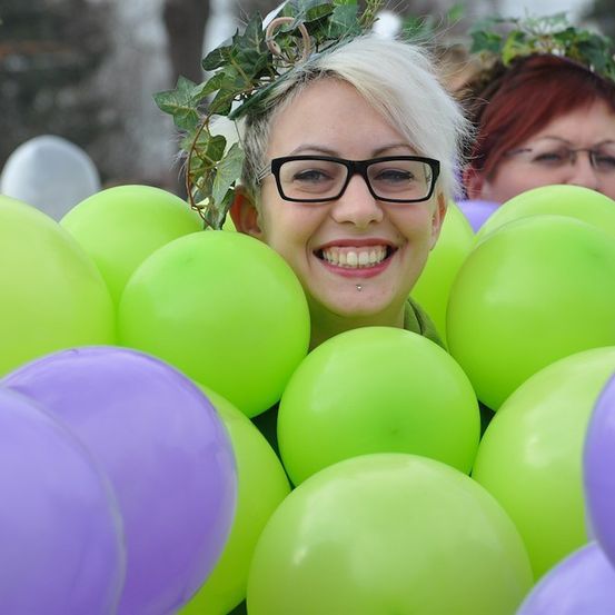 Eine Frau mit blonden Haaren, die eine Brille und eine Krone aus Blättern trägt, ist von grünen und lilafarbenen Luftballons umgeben und lächelt für ein Foto.