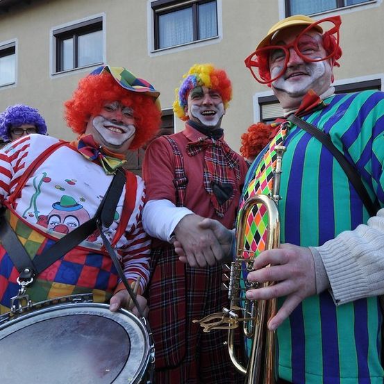 Vier Clowns in bunten Kostümen stehen vor einem Gebäude, einer hält eine Trompete und ein anderer ein Schlagzeug.