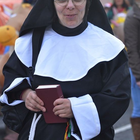 Eine Nonne mit Brille und einem weißen Habit hält ein Buch in den Händen und lächelt in die Kamera.