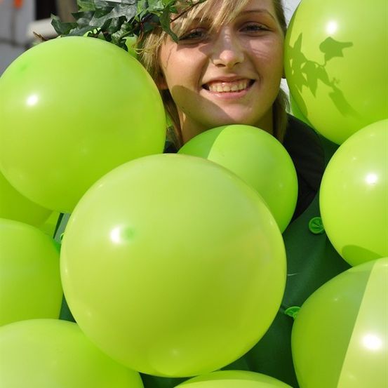 Eine Frau umgeben von grünen Luftballons, mit einem grünen Kranz aus Blättern, lächelt für ein Foto.