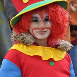 Ein junger Mensch, der als Clown gekleidet ist, trägt eine rote Perücke und Gesichtsbemalung, mit einem roten Hut, der mit einer gelben Blume verziert ist.