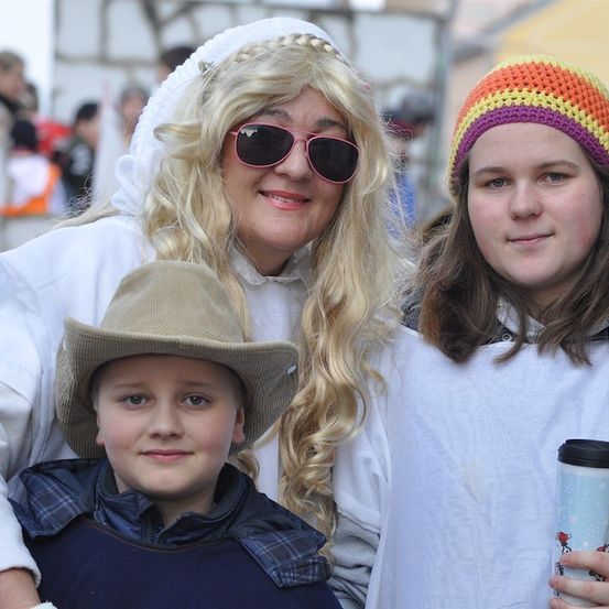 Eine Frau mit blonden Haaren und einem weißen Kopftuch posiert mit einem Jungen in einem Hut und einem Mädchen mit einer bunten Mütze, alle lächeln für die Kamera.