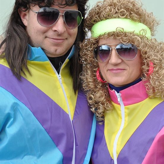 Ein Mann und eine Frau in 1980er-Jahre-Kleidung, beide mit Sonnenbrillen und farbenfrohen Jacken. Der Mann hat lange Haare und einen Schnurrbart, die Frau lockige Haare und eine Stirnband.