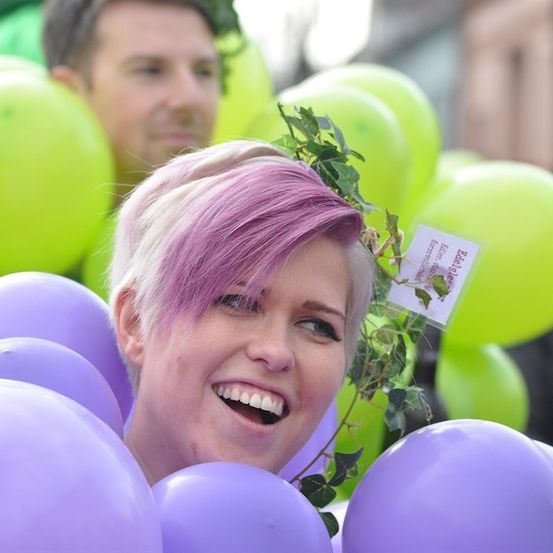 Eine Frau mit rosa Haaren lächelt und ist von lila Luftballons umgeben, mit einem Mann und grünen Luftballons im Hintergrund.