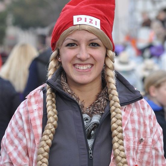 Eine Frau mit langen geflochtenen Haaren, die eine rote Mütze mit dem Wort 'Pizi' trägt und in die Kamera lächelt.