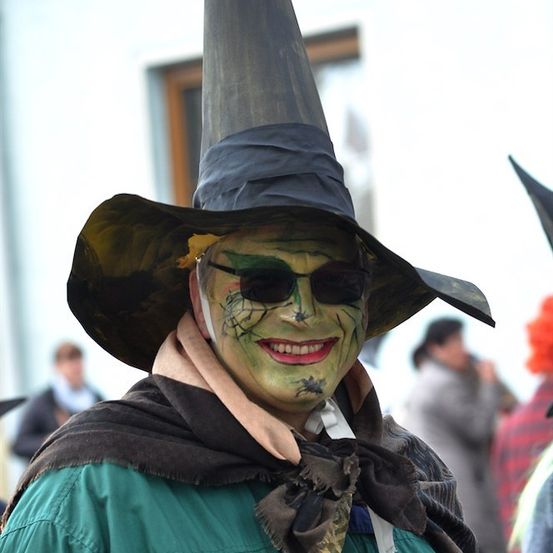 Eine Person, die als Hexe verkleidet ist, mit einem hohen spitzen Hut, grüner Gesichtsbemalung und Sonnenbrille, lächelt in die Kamera.