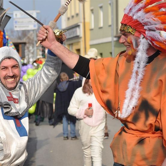 Zwei Männer in Kostümen, einer in einem silbernen Raumanzug und der andere in einem orangefarbenen Indianerhäuptling, halten ein Schwert und eine Glocke, lächeln und posieren für ein Foto. Hinter ihnen gehen Menschen auf der Straße.