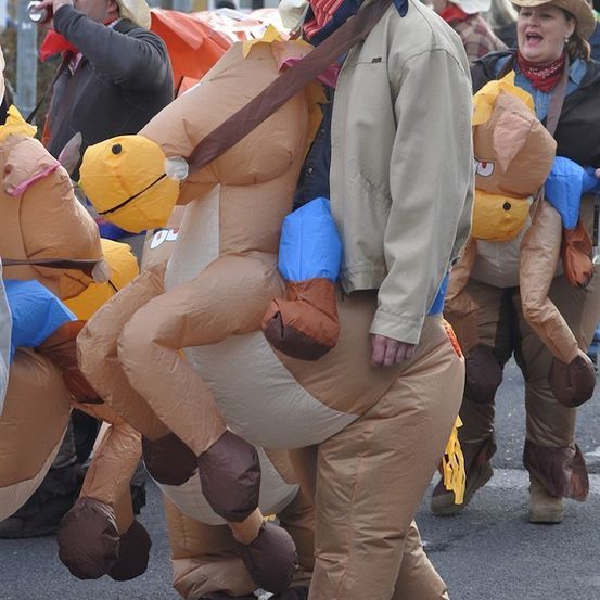 Eine Gruppe von Menschen in Cowboy-Kostümen, eine Person reitet auf einem aufblasbaren Pferd, während andere hinter ihr hergehen. Sie sind in einem Umzug mit Gebäuden im Hintergrund.