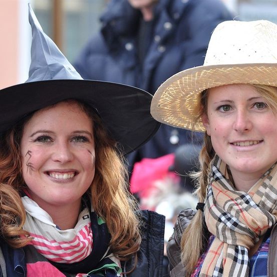 Zwei Frauen in Kostümen, eine trägt einen Hexenhut und die andere einen Strohhut, lächeln für ein Foto.
