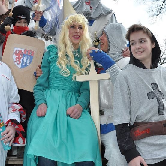 Eine Gruppe von Menschen in mittelalterlichen Kostümen, darunter eine Frau in einem grünen Kleid, die auf einem hölzernen Thron sitzt, mit einem Jungen in einer grauen Kapuzenjacke, der neben ihr lächelt.