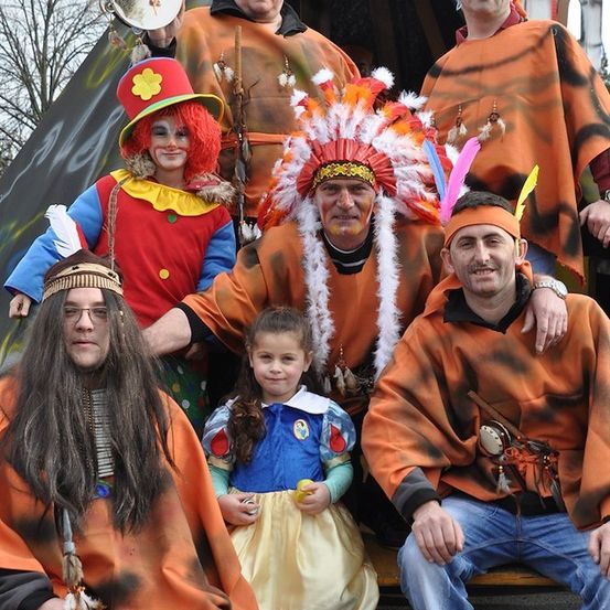 Eine Gruppe von Erwachsenen und einem Kind in verschiedenen Kostümen, darunter ein Clown, eine indianische Kopfbedeckung und ein Schneewittchen-Kleid, posiert für ein Foto.