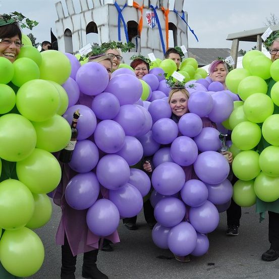 Eine Gruppe von Menschen in Traubenkostümen, die Trauben aus grünen und lila Luftballons halten, lächeln und posieren für ein Foto. Dahinter steht eine burgartige Struktur mit einer Flagge.