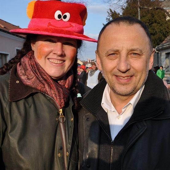 Ein Mann und eine Frau stehen eng beieinander und lächeln für die Kamera. Die Frau trägt einen roten Hut mit Gesicht und eine Schürze. Der Mann trägt einen schwarzen Mantel.