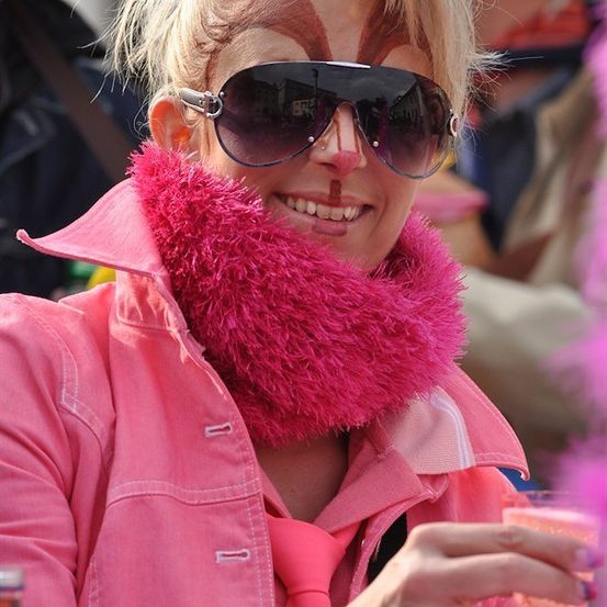 Eine Frau mit pinkem Gesichts-Make-up und einem rosa Pelzschal lächelt und hält ein Getränk bei einer Veranstaltung im Freien.