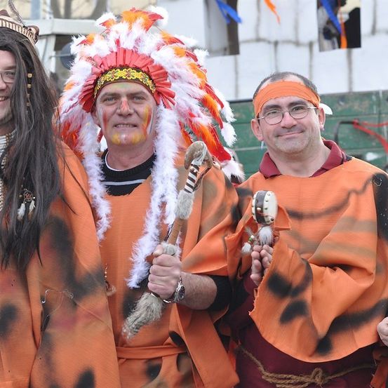 Vier Männer in orange und schwarzen Kostümen, möglicherweise für ein Festival, lächeln und posieren für ein Foto. Ein Mann trägt eine Kopfbedeckung, ein anderer hat Gesichtsbemalung, und sie alle halten Requisiten.