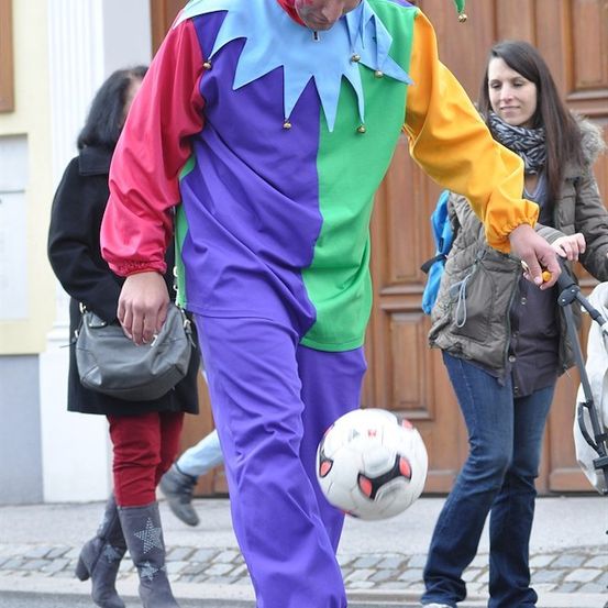 Ein Mann in einem farbenfrohen Narrenkostüm balanciert einen Fußball auf der Straße. Hinter ihm schiebt eine Frau einen Kinderwagen und eine weitere Frau geht vorbei.