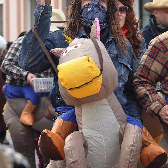 Eine Person in einer Jeansjacke reitet auf einem großen aufblasbaren Pferd bei einer Parade und hält eine Pistole. Eine Frau mit Maske und eine weitere Person in einem Cowboyhut sind in der Nähe.
