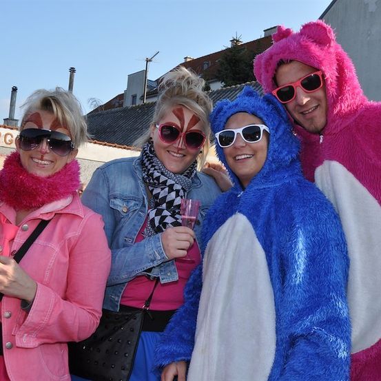 Vier Personen in rosa und blauen Kostümen, mit Sonnenbrillen und Getränken in der Hand, posieren für ein Foto. Hinter ihnen steht ein Gebäude mit Dach und Schornstein.