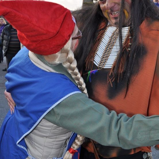 Eine Person in einem roten Hut und blauem Kleid umarmt eine Person in einem Indianerkostüm mit Gesichtsbemalung.