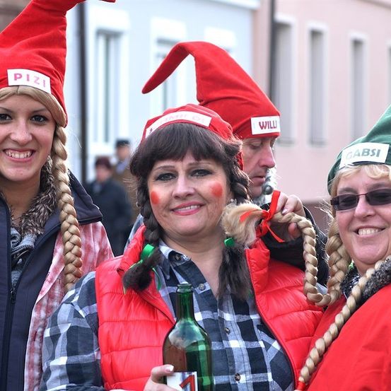 Vier Frauen in roten und grünen Elfenhüten, lächelnd und eine Flasche haltend, mit einem Gebäude im Hintergrund.