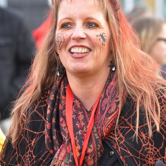 Eine Frau mit langen blonden Haaren und Gesichtsbemalung lächelt in die Kamera. Sie trägt einen gemusterten Schal und eine rote Kordel.