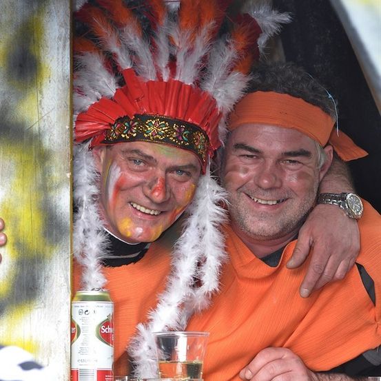 Zwei Männer in orangefarbener Kleidung, einer mit Kopfschmuck, lächeln und posieren für ein Foto mit einer Bierdose und einem Glas Flüssigkeit vor ihnen.