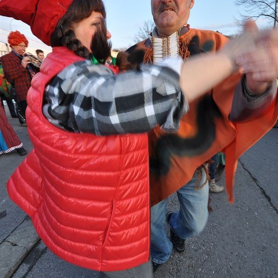 Ein Mann und eine Frau in festlicher Kleidung tanzen auf der Straße. Die Frau trägt einen roten Hut und Weste, der Mann hat einen Schnurrbart und ein orangefarbenes Outfit.
